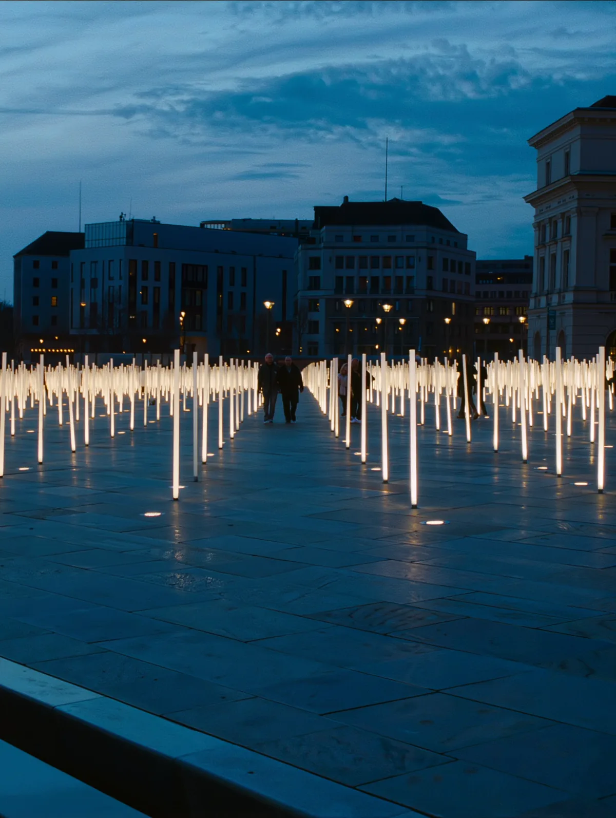 Outdoor public art plaza at dusk with light columns brightening around pedestrians