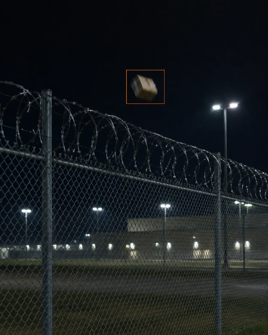 Package thrown over a razor-wire fence at night outlined in orange