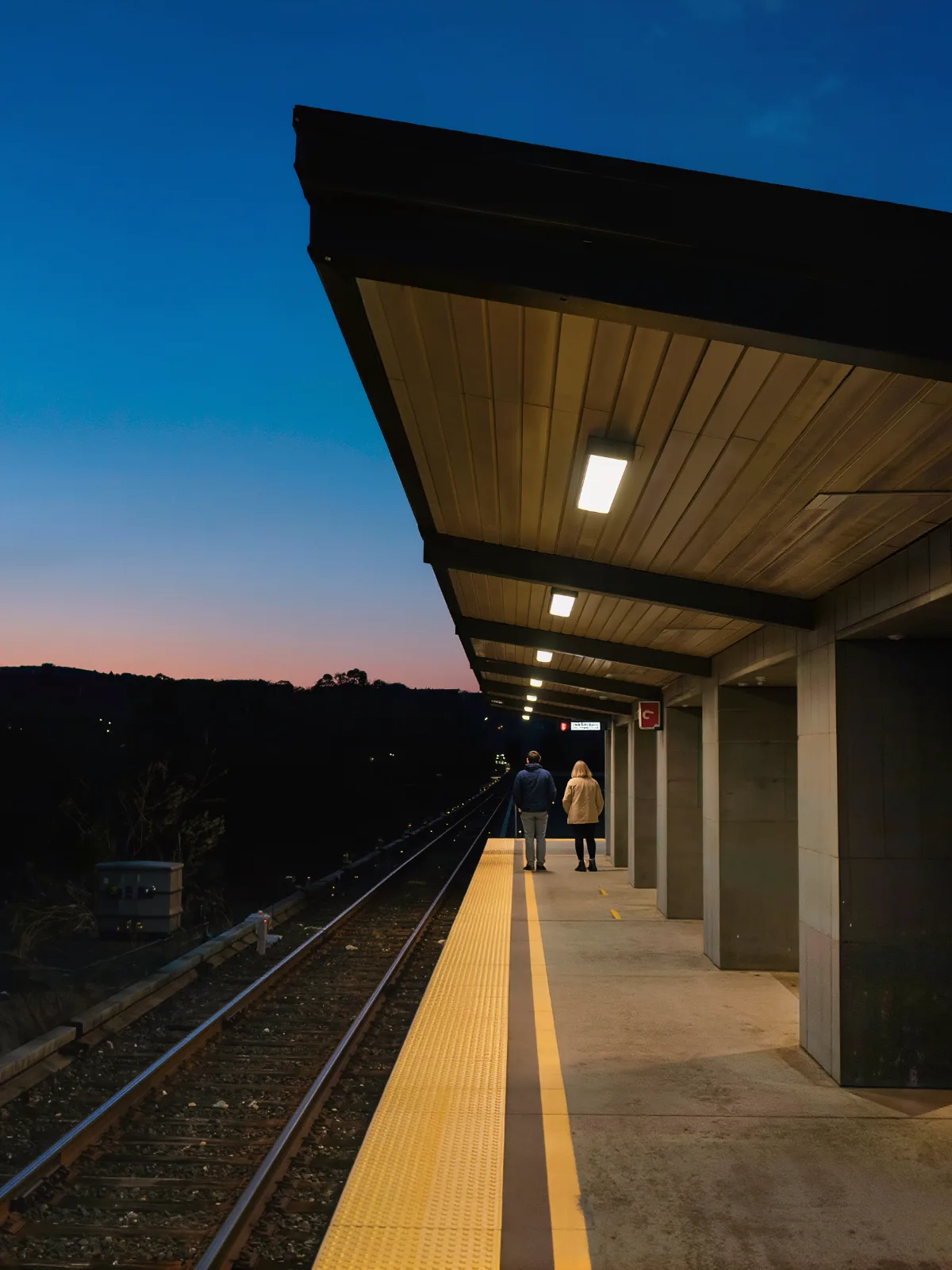 Commuter rail platform at dusk with a yellow safety line and two passengers